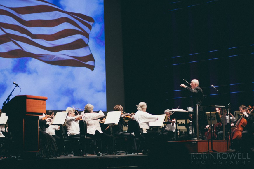 The maestro leads the symphony while an American flag flies behind on the screen